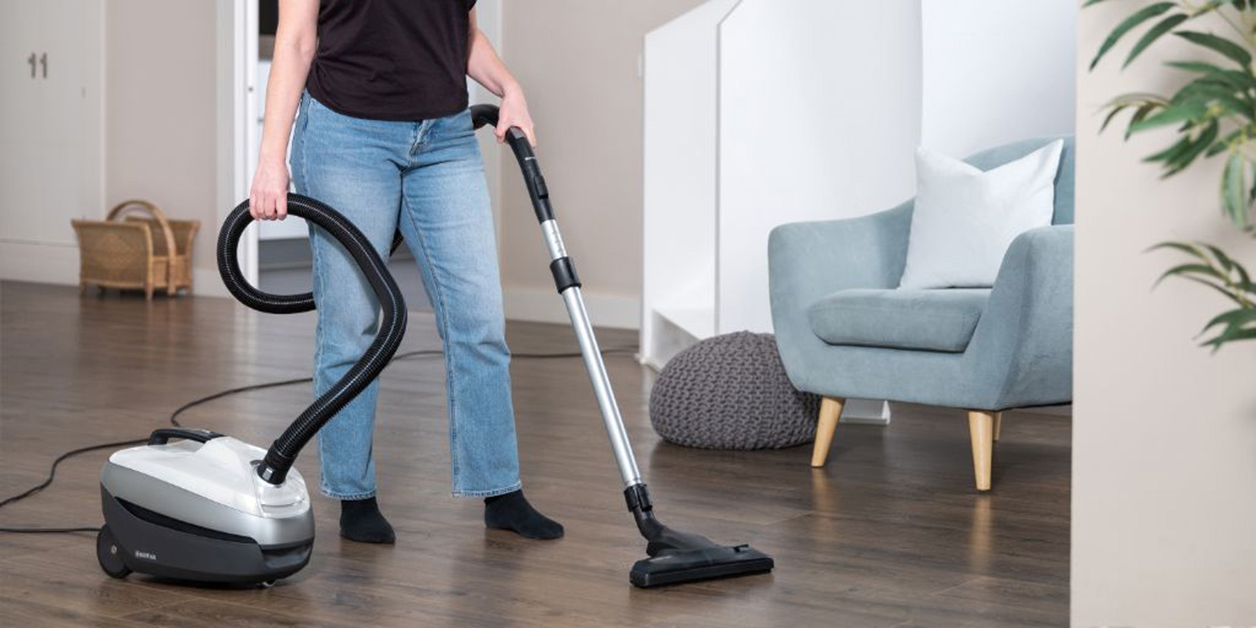 Person vacuuming hardwood floor in bright, tidy living room with blue armchair and green plant.