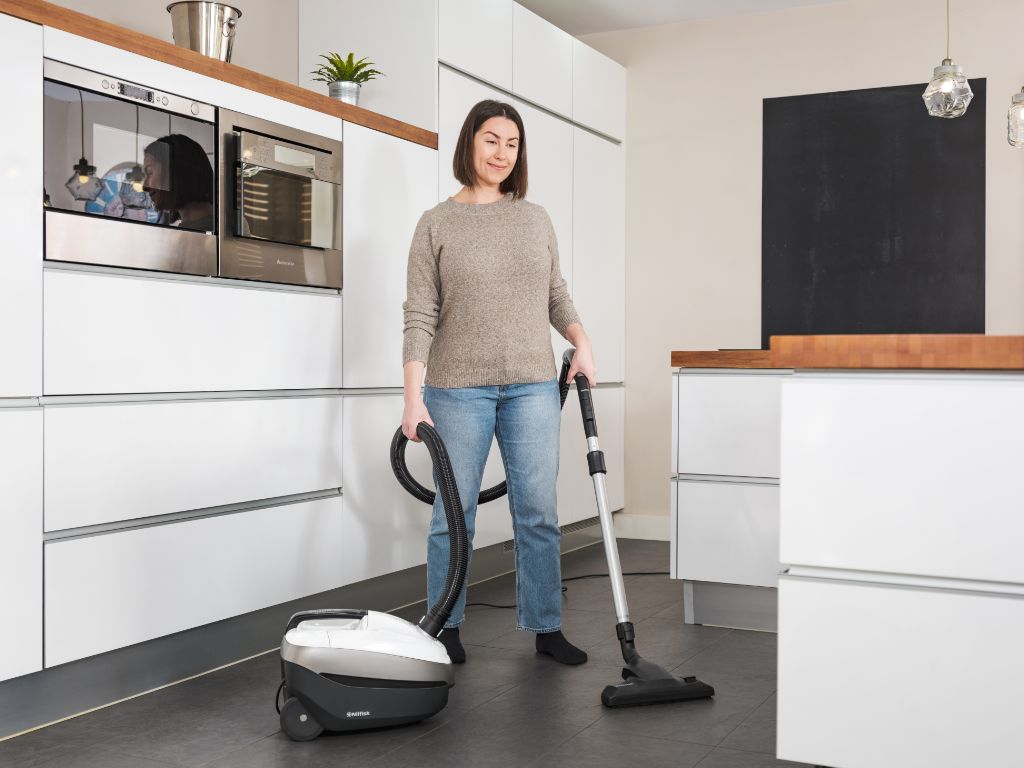 Woman smiling while using a traditional canister vacuum in a modern, bright kitchen with white cabinets and dark flooring.