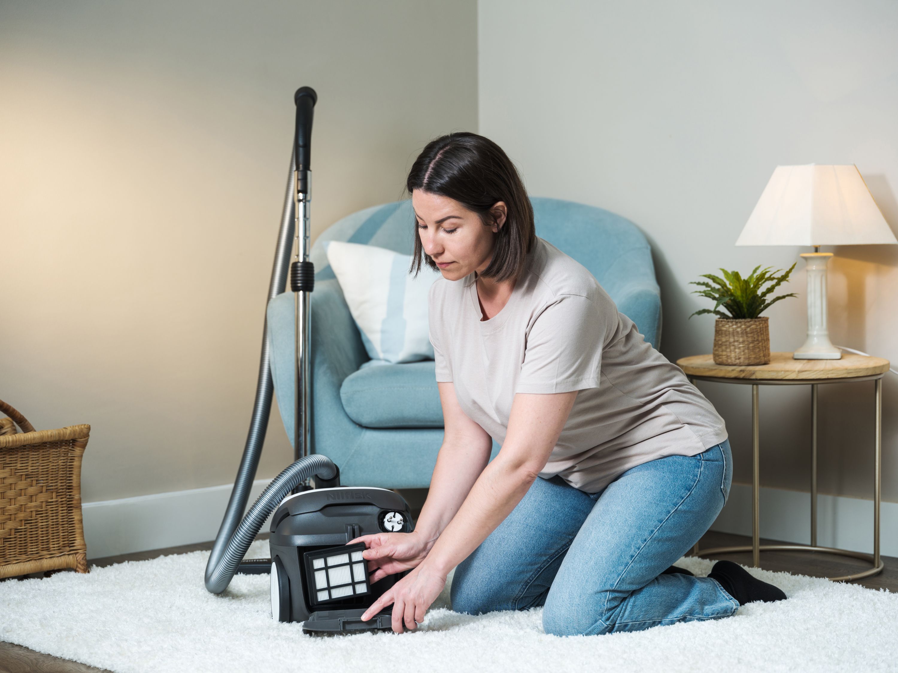 Woman kneeling on white rug, carefully removing vacuum filter from canister in cozy living room.
