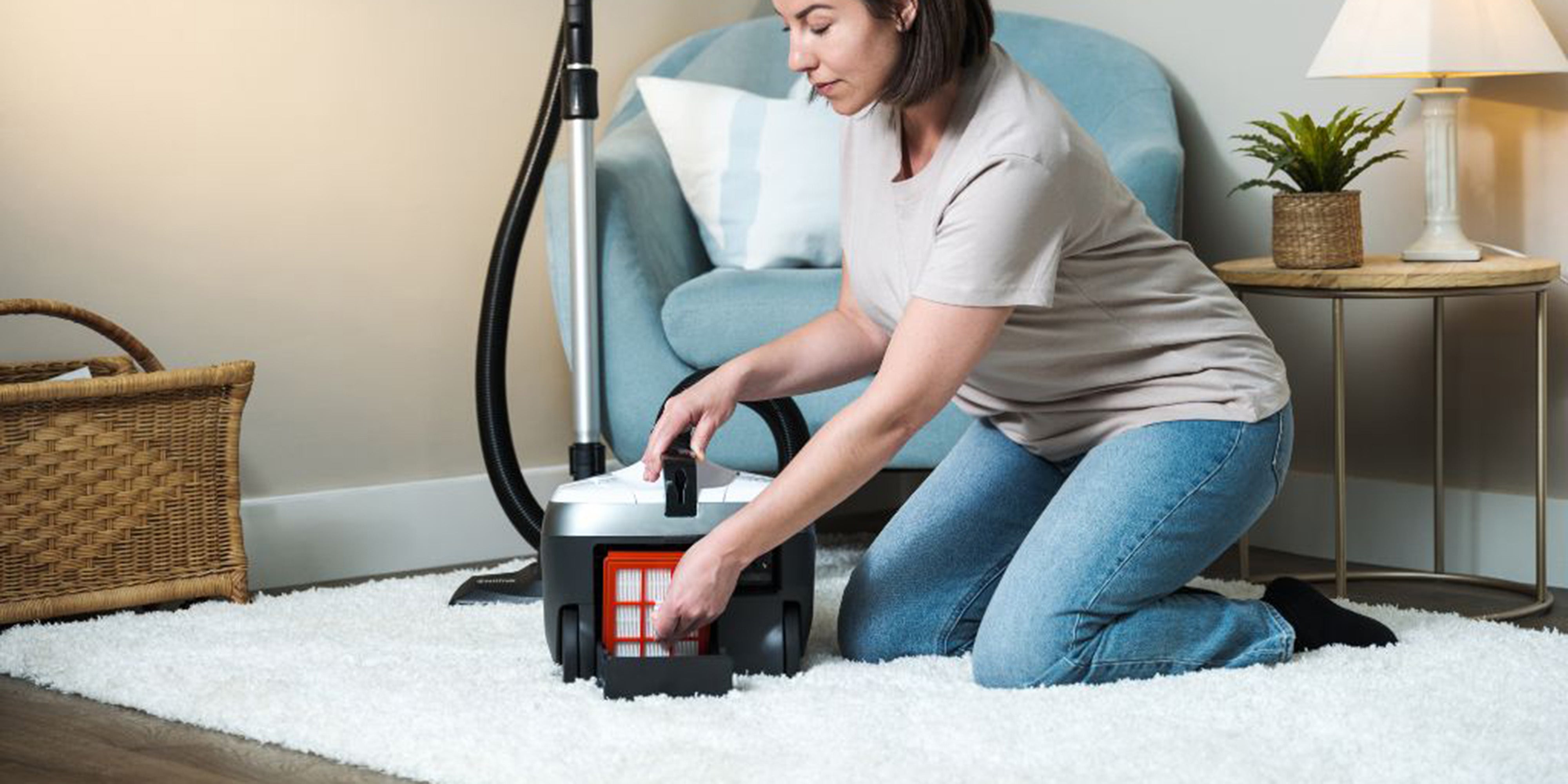 Woman kneeling on white rug, carefully removing filter from upright vacuum cleaner in cozy living room.