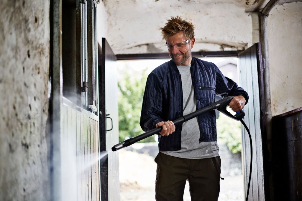 Man using high-pressure washer to clean horse stable walls, wearing safety glasses and smiling.
