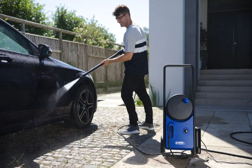 Man using blue pressure washer to clean black car in sunny driveway, water spraying from nozzle.