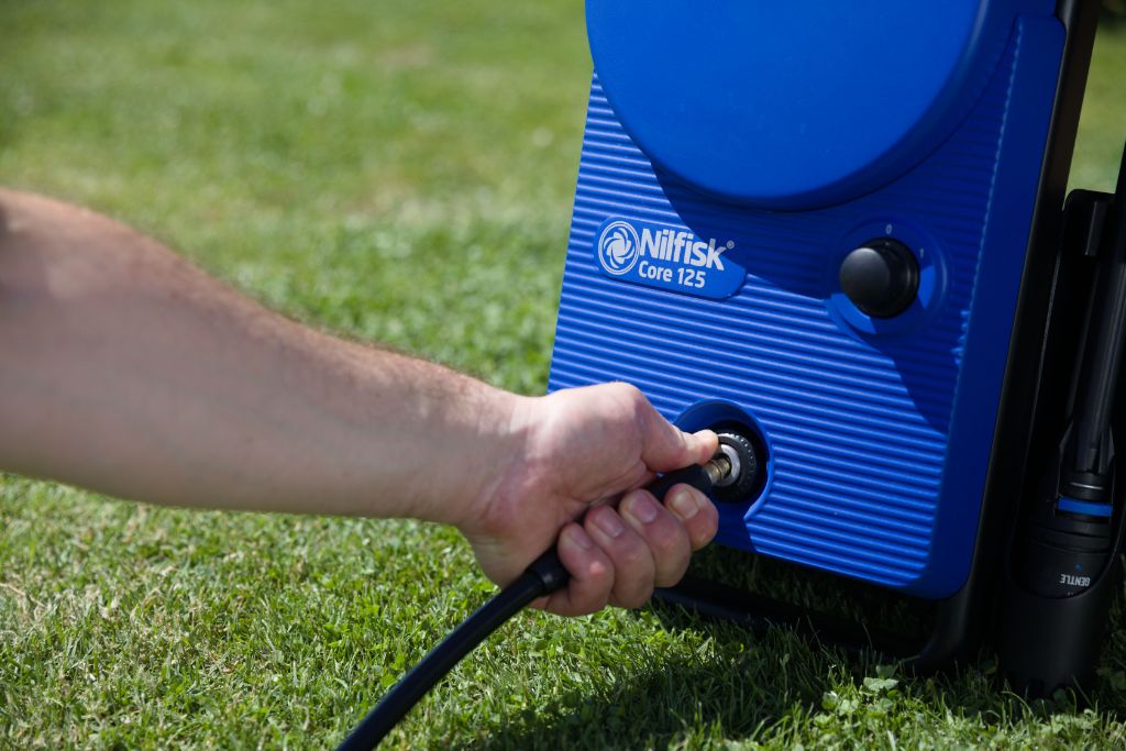 Hand connecting hose to blue Nilfisk Core 125 pressure washer on green lawn, demonstrating maintenance step.
