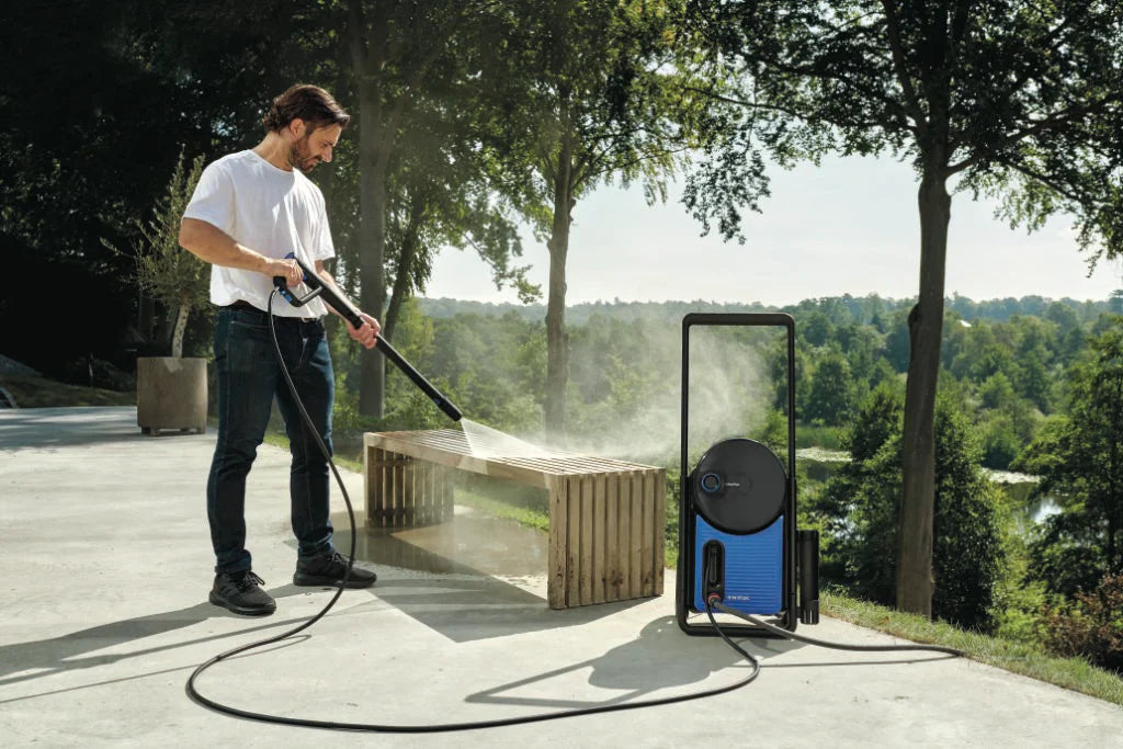 Man cleaning wooden bench with blue pressure washer in sunny garden, mist spraying, trees in background.