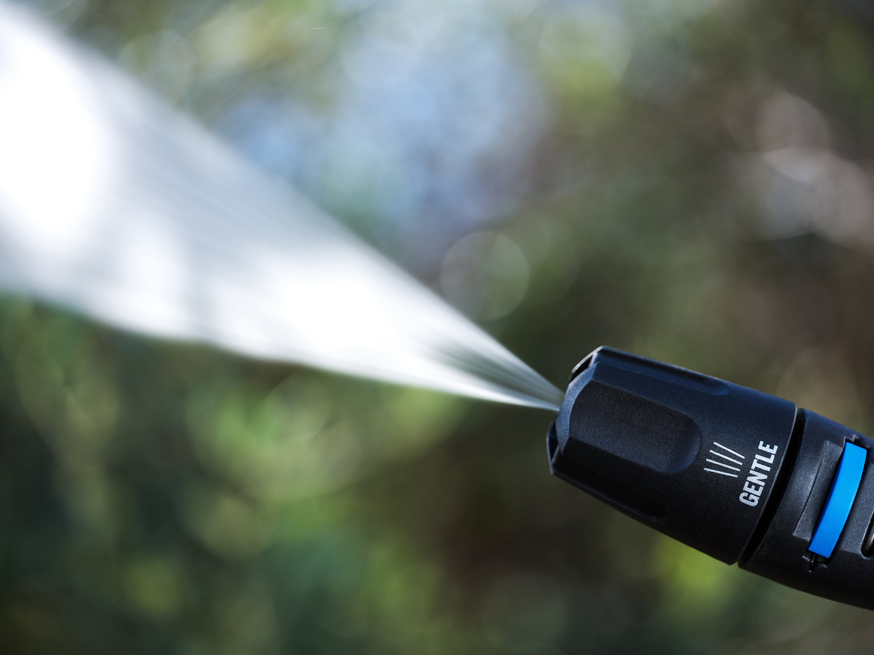 Close-up of pressure washer nozzle spraying water in gentle mode, set against blurred green garden backdrop.