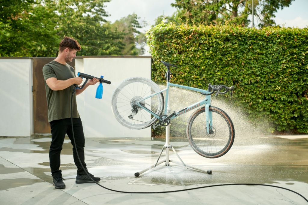 Man cleaning light blue bike with pressure washer in backyard, water spraying, green hedge background.