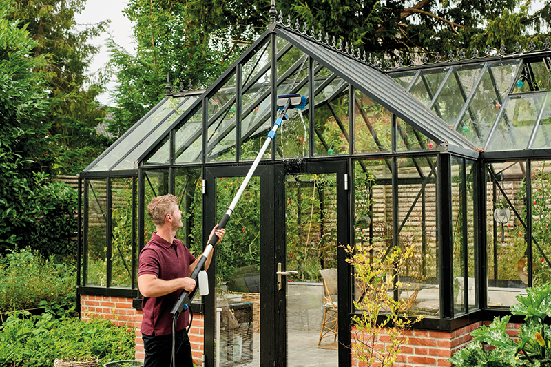 Man uses extendable cleaning lance to wash greenhouse glass, reaching high panels with ease in a lush garden.