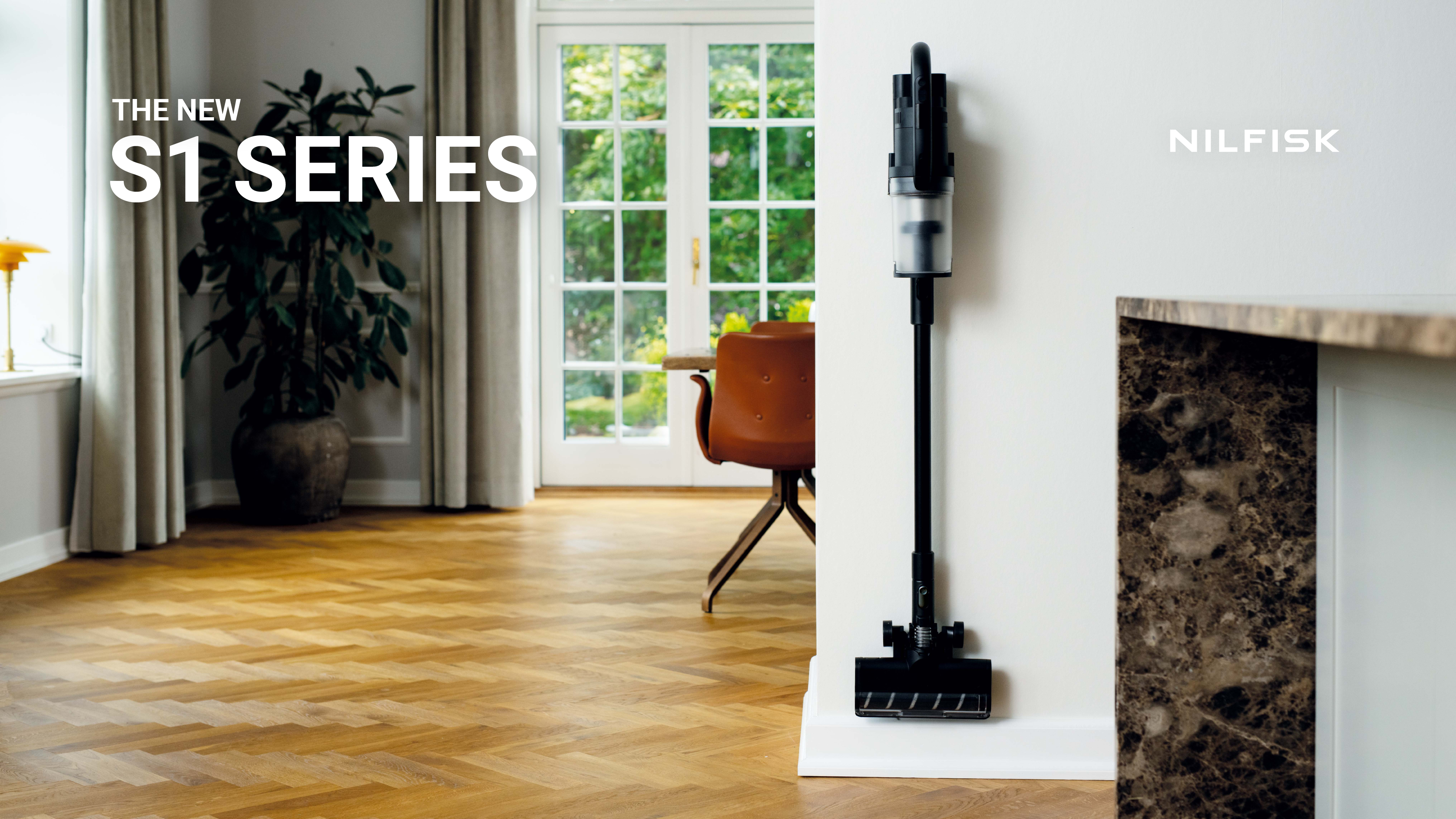 Sleek black cordless vacuum mounted on white wall in bright, modern living room with wooden floors and greenery.
