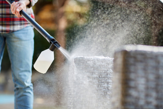 Person using high-pressure washer with detergent attachment to clean outdoor brick surface, creating misty spray.