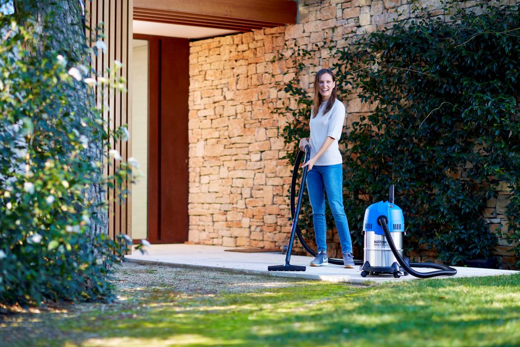 Woman smiles while using wet/dry vacuum outdoors near stone house, clearing debris from patio.