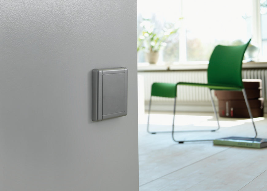 Modern silver light switch on gray wall, bright living room with green chair and sunlight streaming through window.