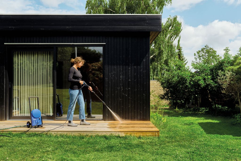 Woman cleaning wooden terrace with pressure washer on sunny day, modern house in background