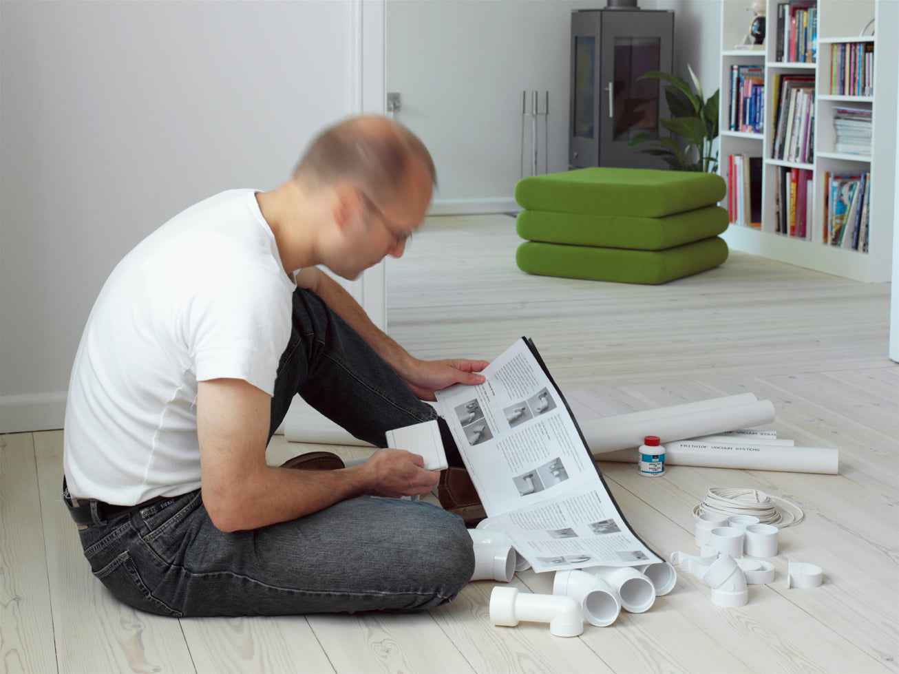 Man installing central vacuum system, reading manual with white pipes and fittings on wooden floor in bright living room.