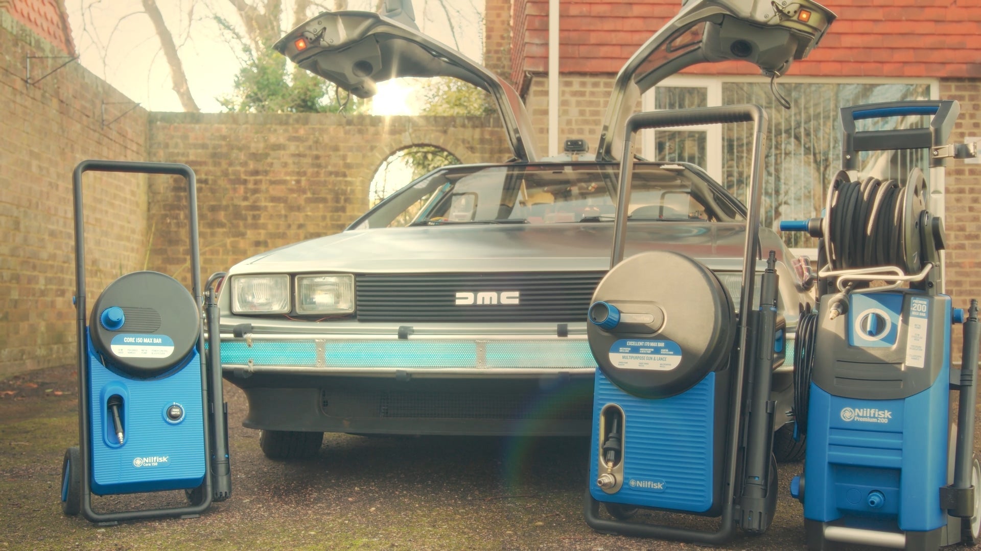 Shiny DeLorean with gull-wing doors flanked by blue Nilfisk pressure washers in a sunlit driveway, ready for cleaning.
