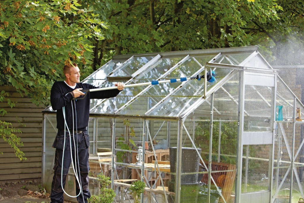 Man cleaning greenhouse glass with high-pressure washer in sunny garden, water spraying from nozzle