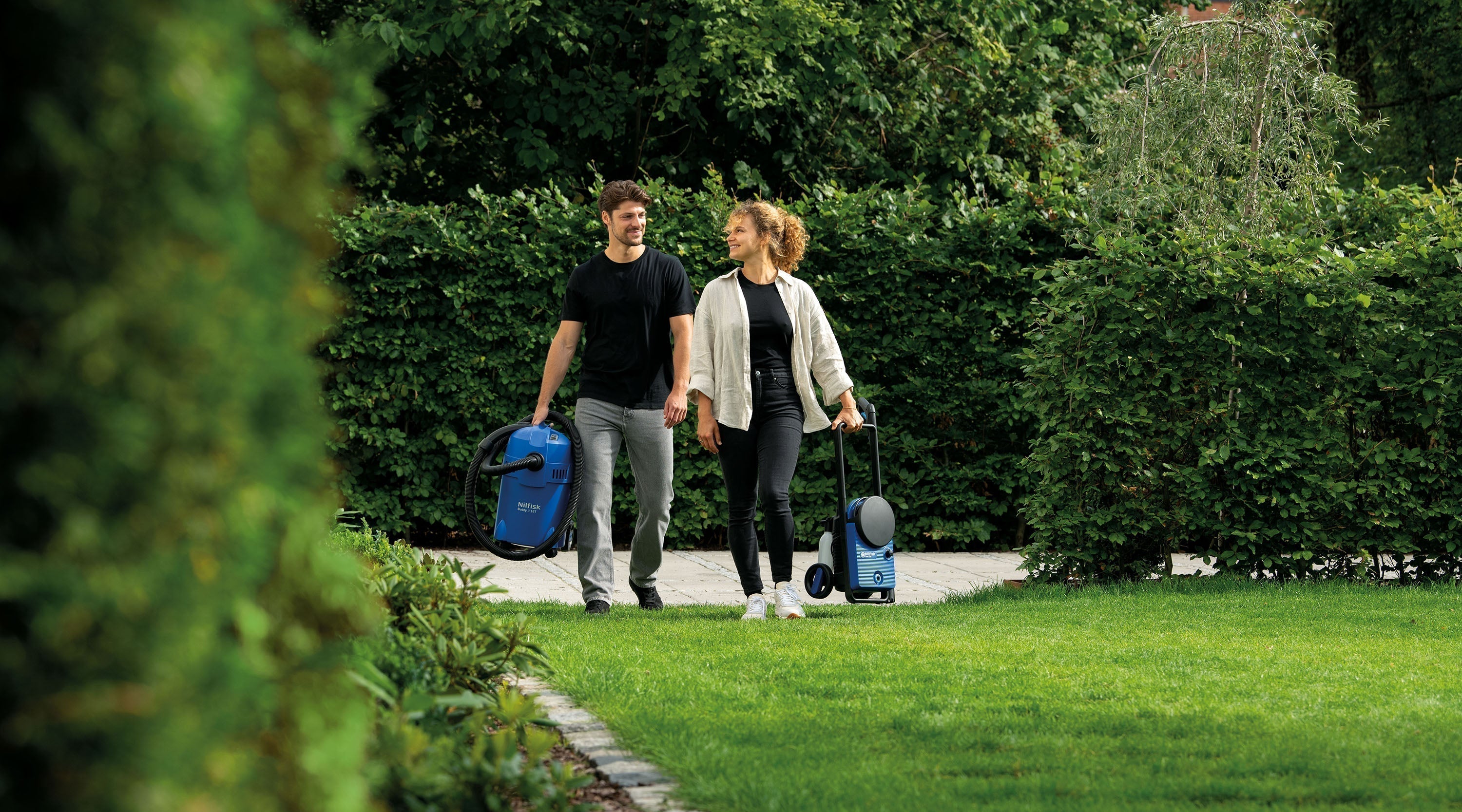 Couple walking in garden with blue pressure washer and wet/dry vacuum, ready to clean artificial turf.