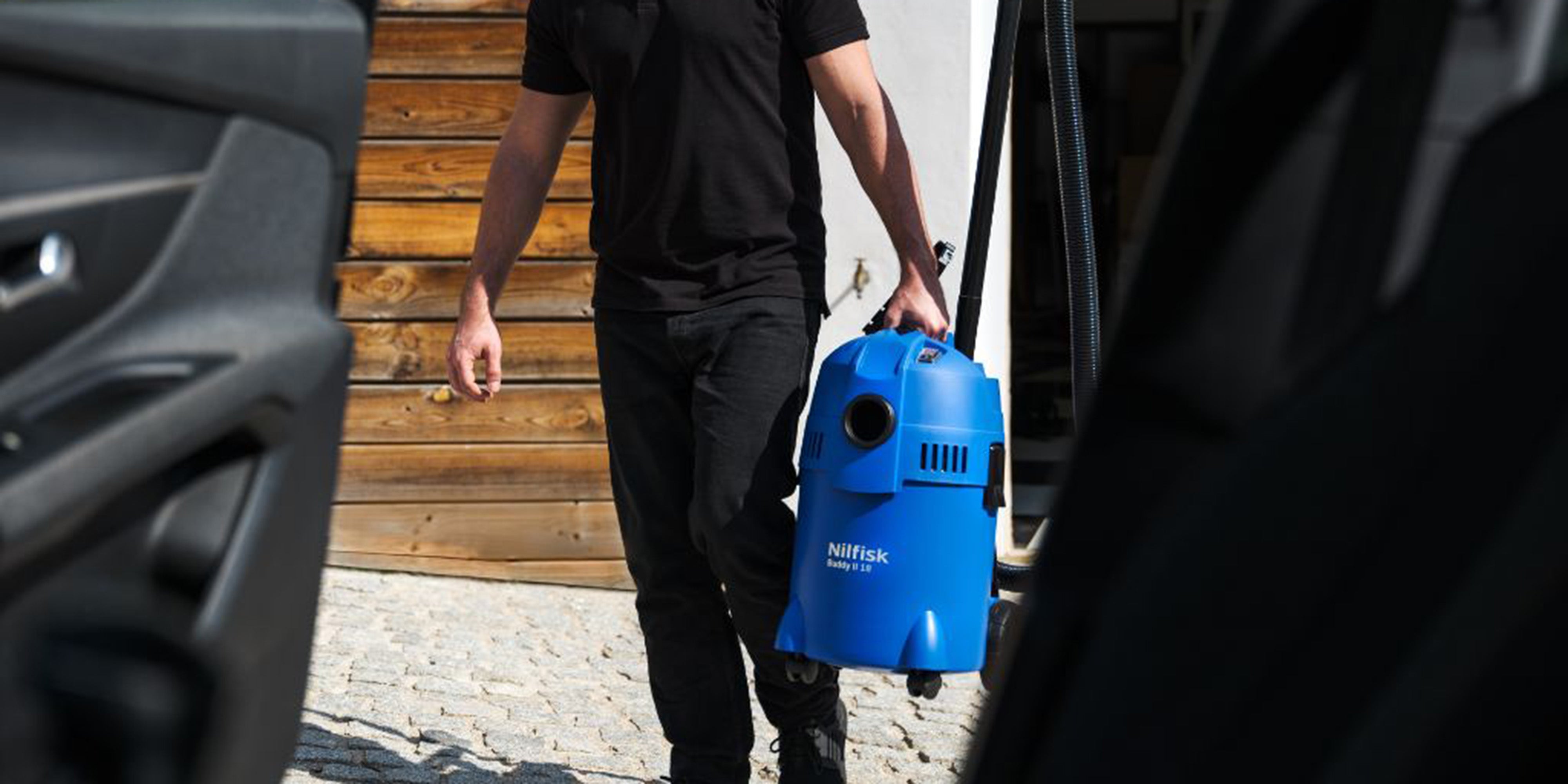 Man in black outfit pushes blue Nilfisk vacuum between cars, preparing for car interior cleaning.