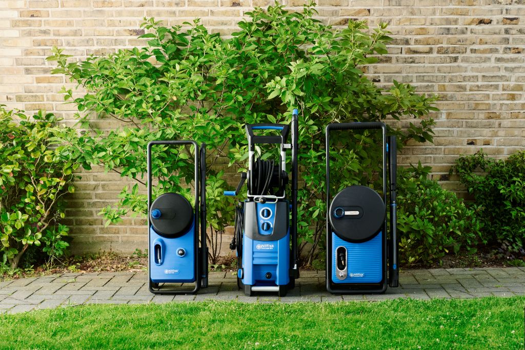 Three blue high-pressure washers lined up on a patio, ready for outdoor cleaning, set against a brick wall and green shrubs.