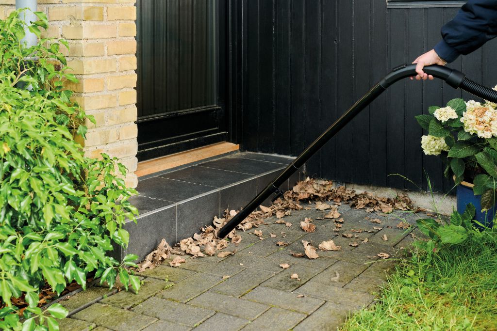 Person using wet/dry vacuum to clean fallen leaves from stone patio near house entrance, surrounded by greenery and flowers.