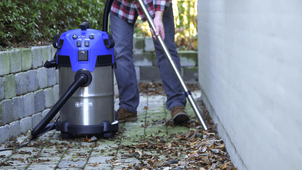 Person using blue and silver wet/dry vacuum to clean leaves from stone patio, outdoor maintenance scene.