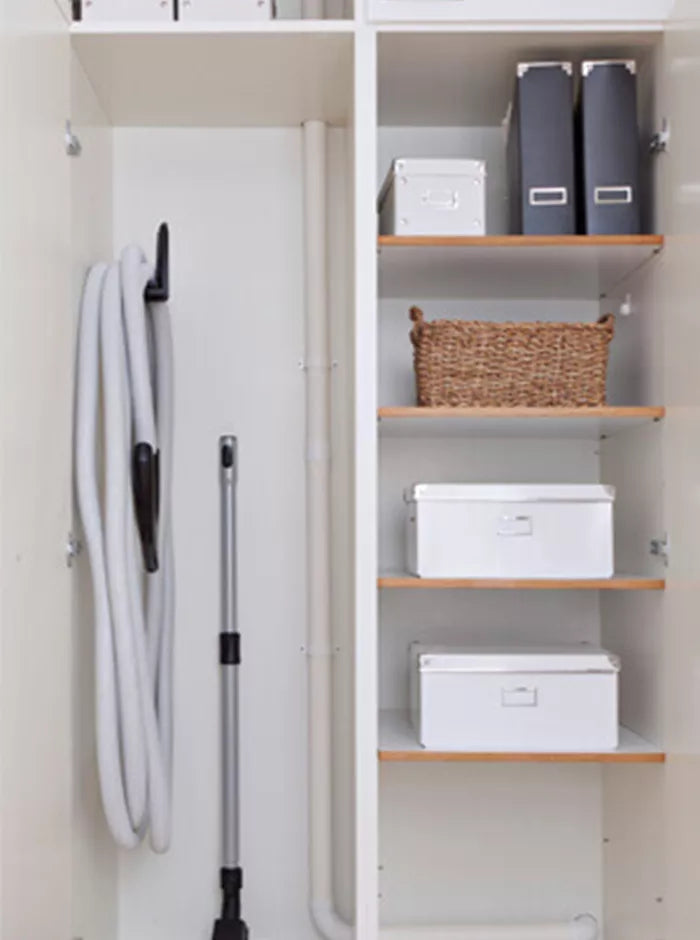 Neatly organized closet with vacuum hose hung on hook, shelves holding white boxes, folders, and a wicker basket.