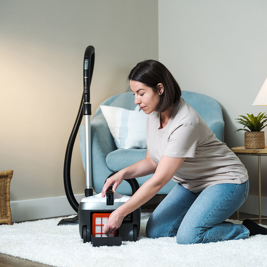 Modern canister vacuum in sleek black and silver, woman easily emptying dust container on plush white rug in bright, cozy living room.