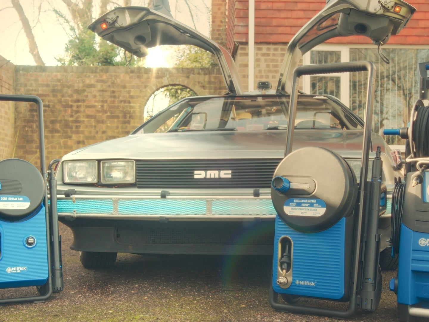 Shiny DeLorean with gull-wing doors flanked by blue Nilfisk pressure washers in a sunlit driveway, ready for cleaning.