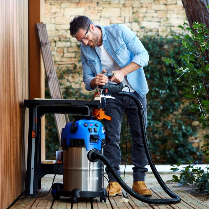Man in blue shirt and glasses uses power tool on wood, connected to blue and silver shop vacuum on deck.