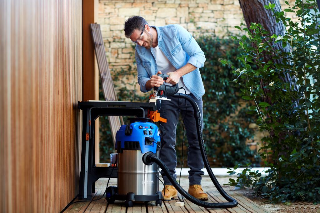 Man in blue shirt and glasses uses power tool on wood, connected to blue and silver shop vacuum on deck.