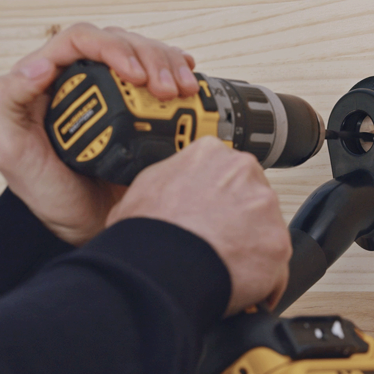 Hands using yellow and black cordless drill to secure black wall mount to light wood surface.