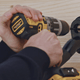 Hands using yellow and black cordless drill to secure black wall mount to light wood surface.