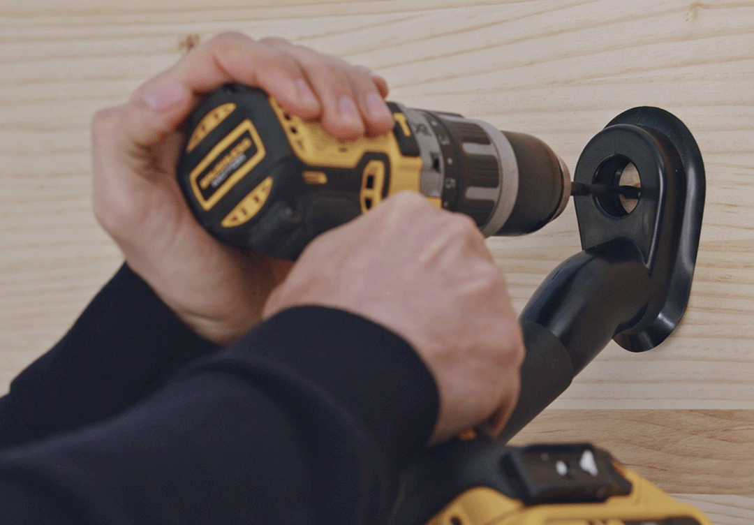 Hands using yellow and black cordless drill to secure black wall mount to light wood surface.
