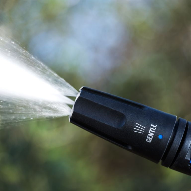 Black power washer lance with blue accents by Nilfisk, emitting a spray of water on a blurred green background.