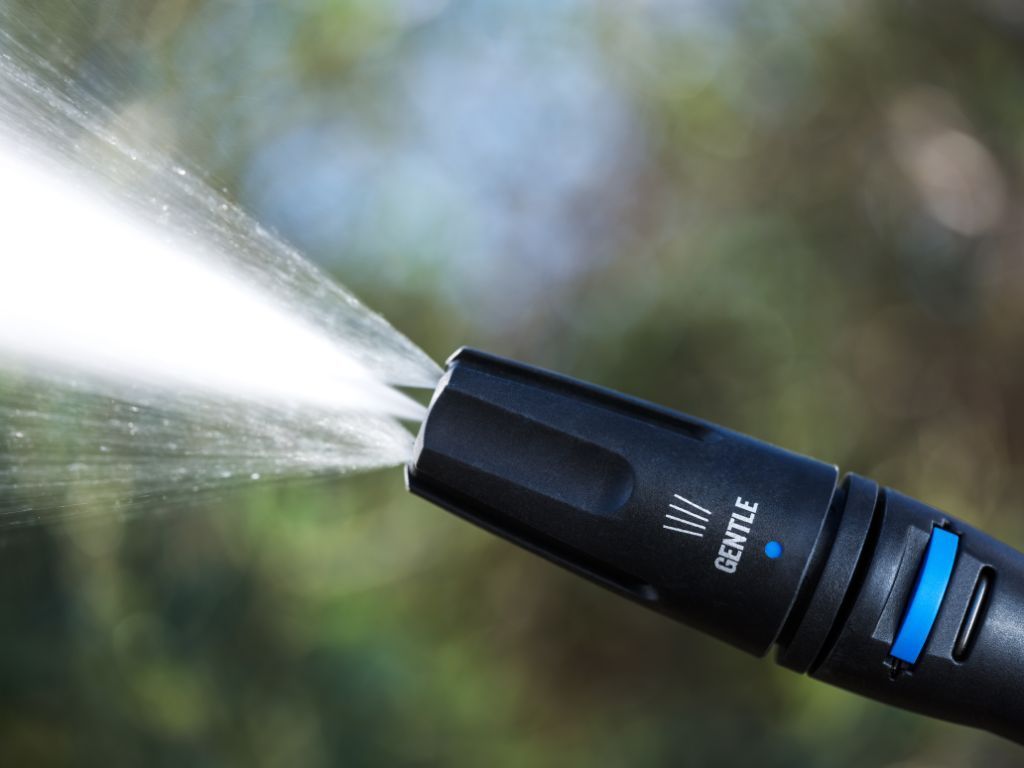 Black power washer lance with blue accents by Nilfisk, emitting a spray of water on a blurred green background.